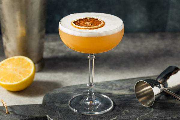 Whiskey Sour in a coupe with a white foam cap and dried citrus slice, beside a lemon and steel bar tools.