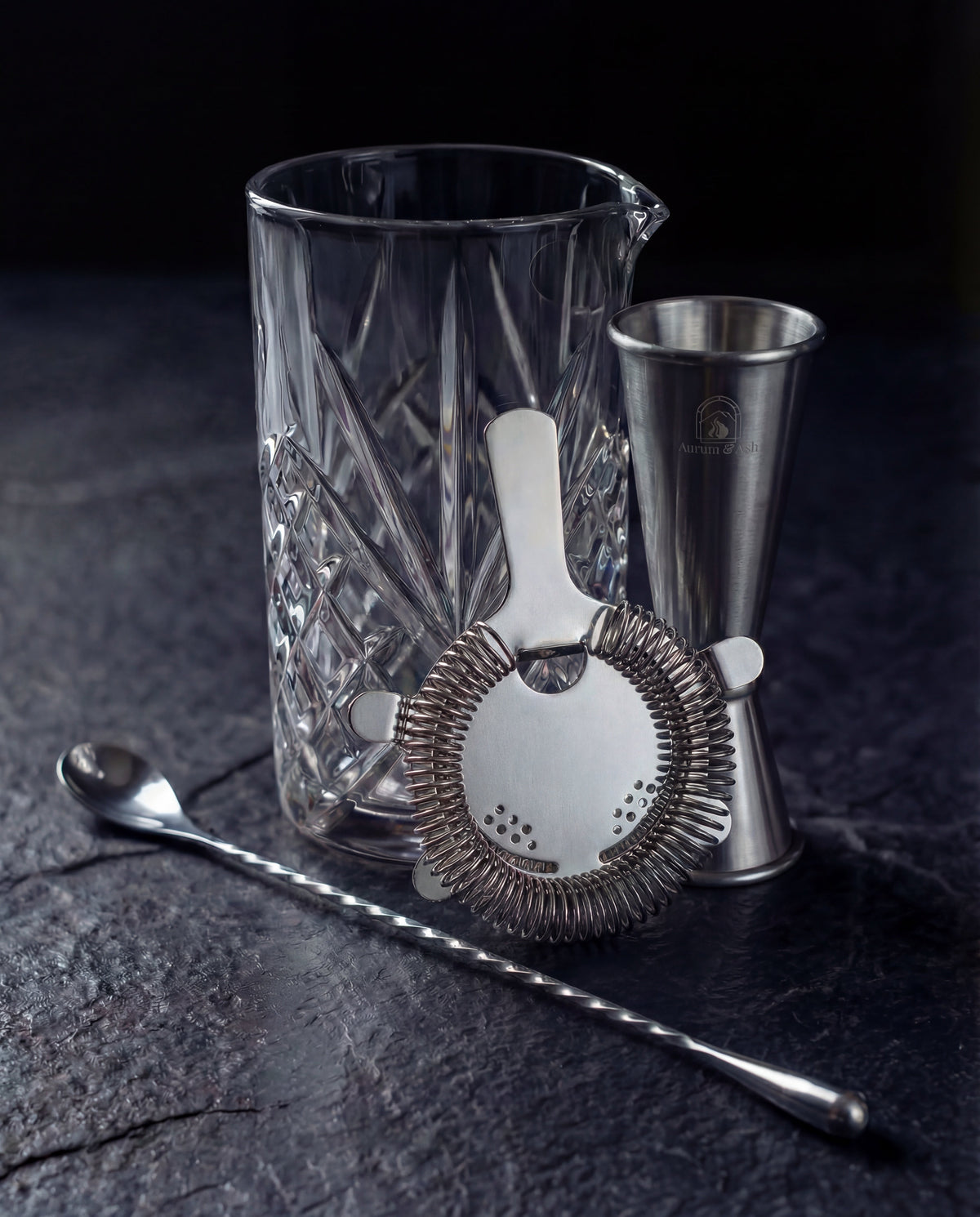 The Stirred Set: Crystal Mixing Glass with the Hawthorne Strainer, Jigger, and Bar Spoon on a dark slate surface.