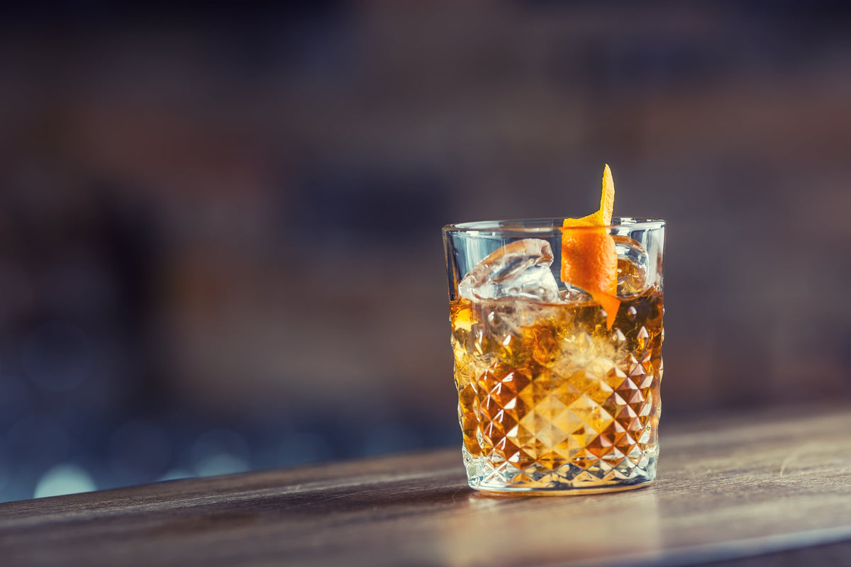 Amber drink with ice and orange peel in a cut-glass tumbler on a wooden counter.