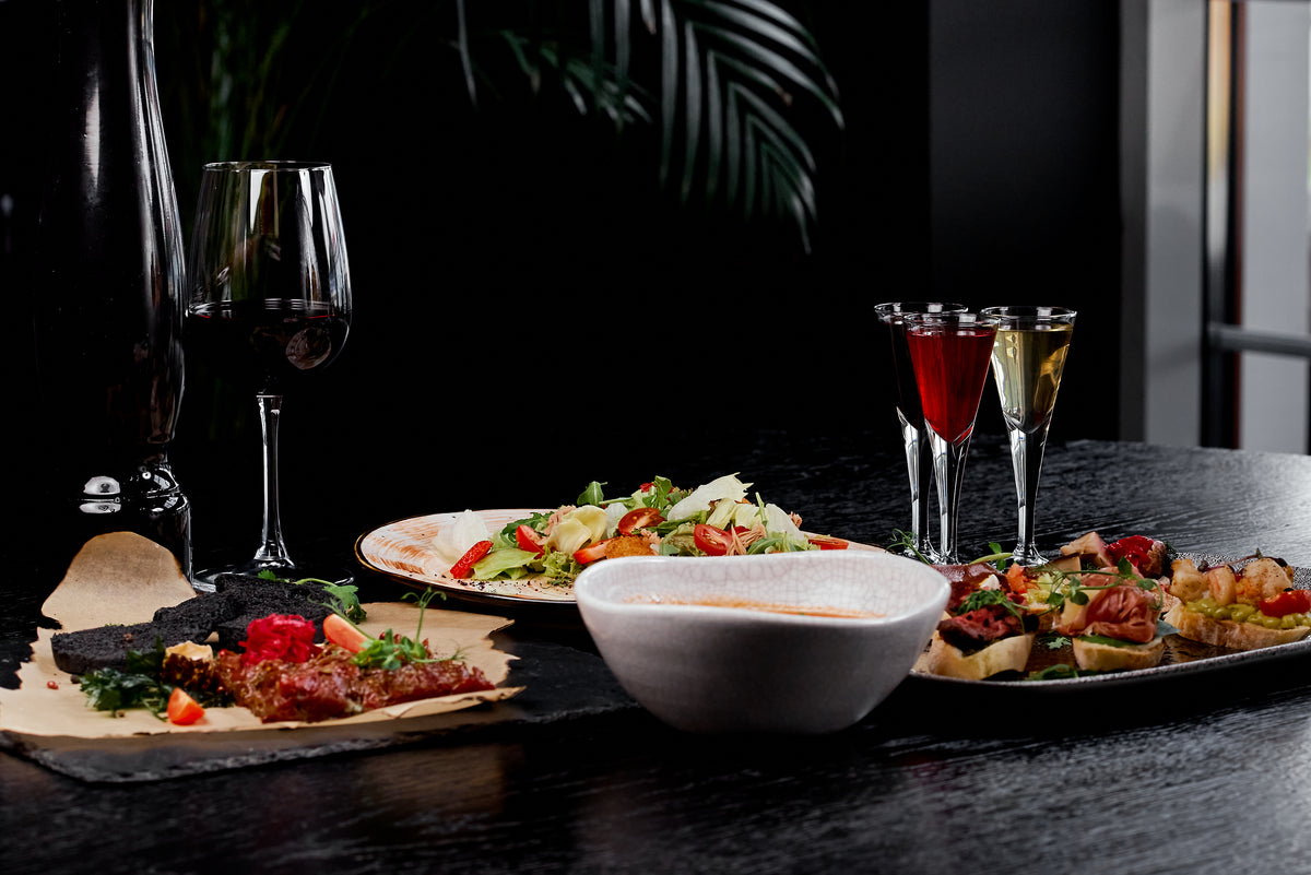 Dinner table set with wine, cordials in stemmed glasses, charcuterie, salad, and bruschetta on dark stone.